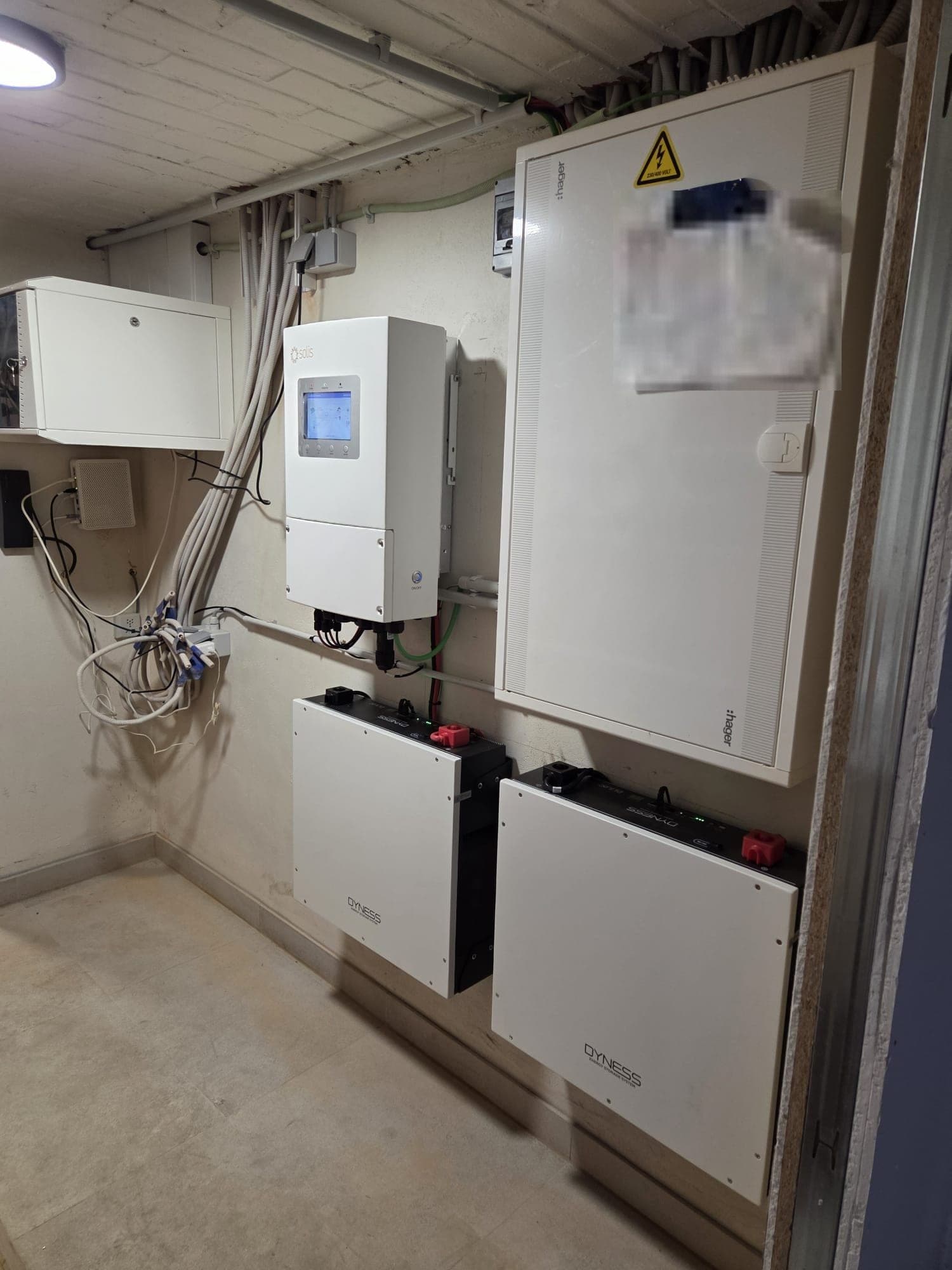 Un onduleur Solis blanc au-dessus de deux modules de batterie Dyness fixés au mur, à côté d'un tableau électrique Hager