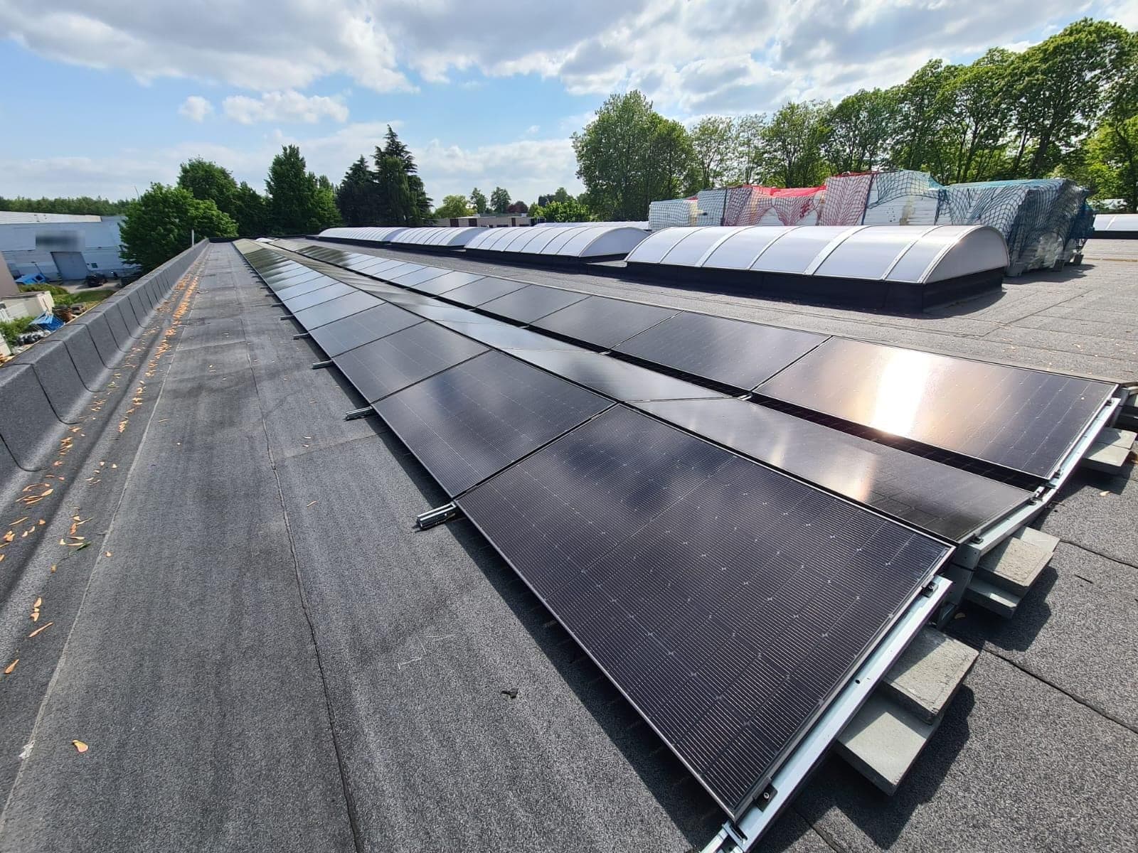 Longues rangées de panneaux solaires noirs sur des supports bas lestés, sur un toit plat commercial, avec des coupoles d'éclairage et des arbres à l'arrière