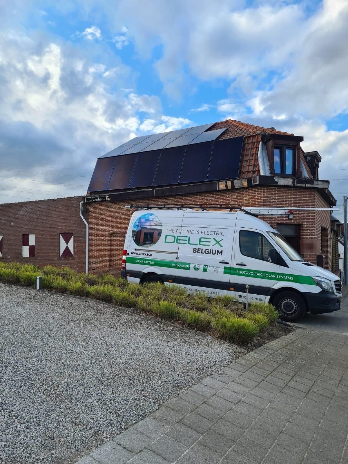 Maison d'angle en briques au toit mansardé couvert de panneaux solaires full black, la camionnette blanche DELEX garée devant